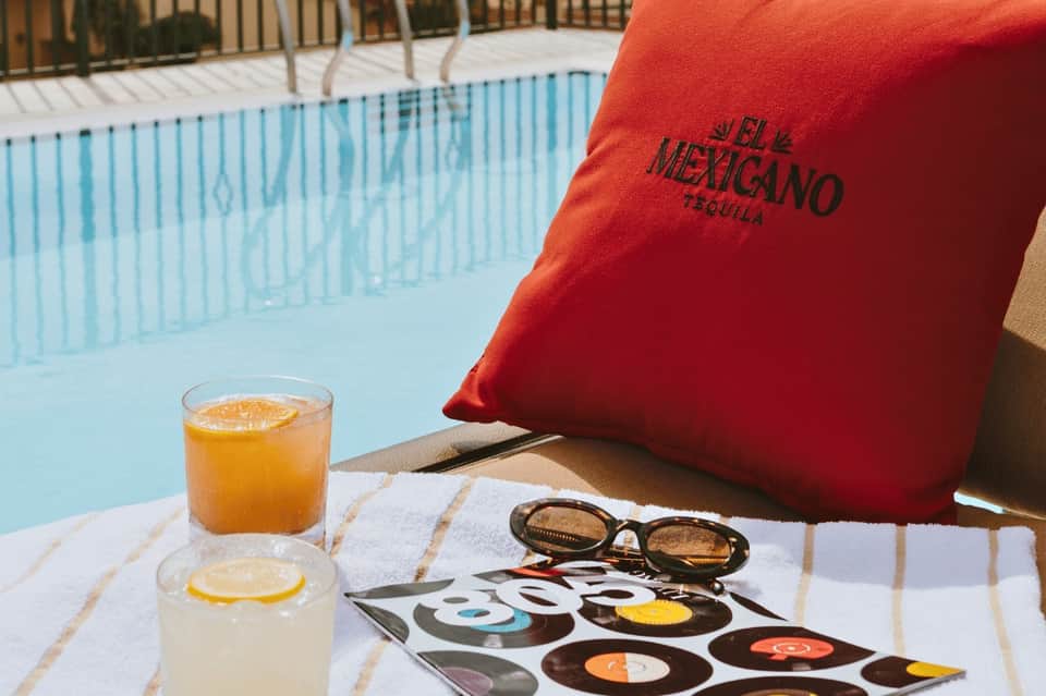 Poolside view with red pillow marked 'El Mexicano Tequila,' orange juice, and sunglasses on table