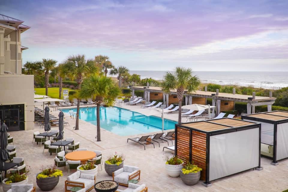 Beachfront pool complex at dusk with palm trees, cabanas, fire pit, lounge chairs, and ocean horizon