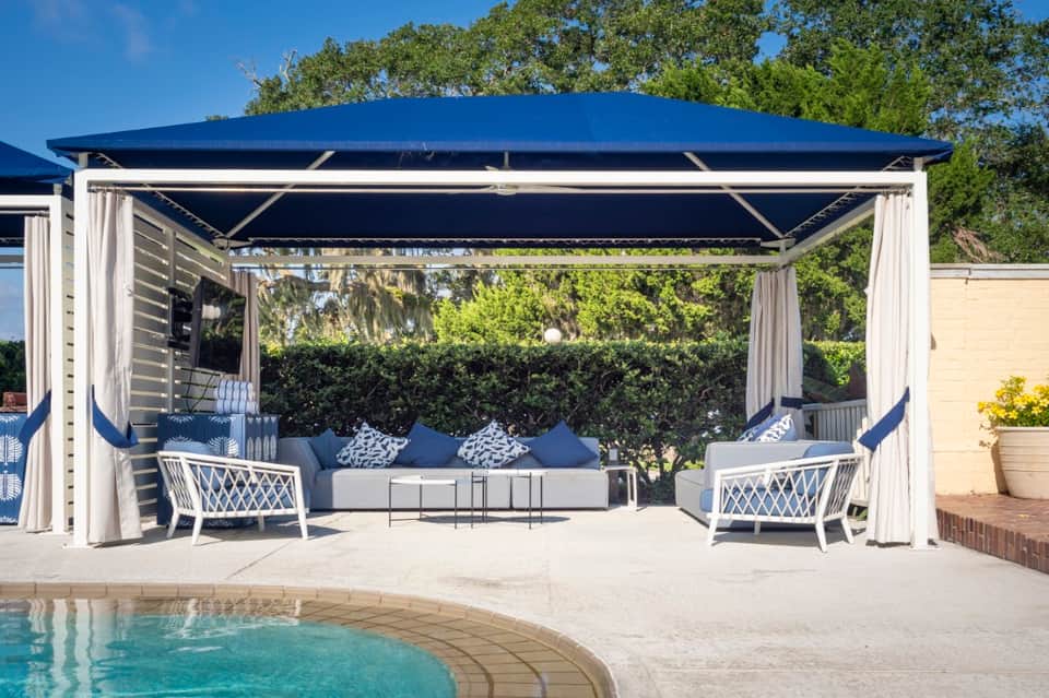 Poolside cabana with retractable awning, white lounge seating, curtains, and integrated pool in background
