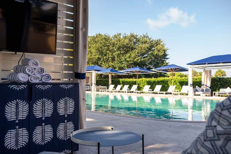 Resort pool area with blue umbrellas, white loungers, decorative storage cabinet, and landscaping