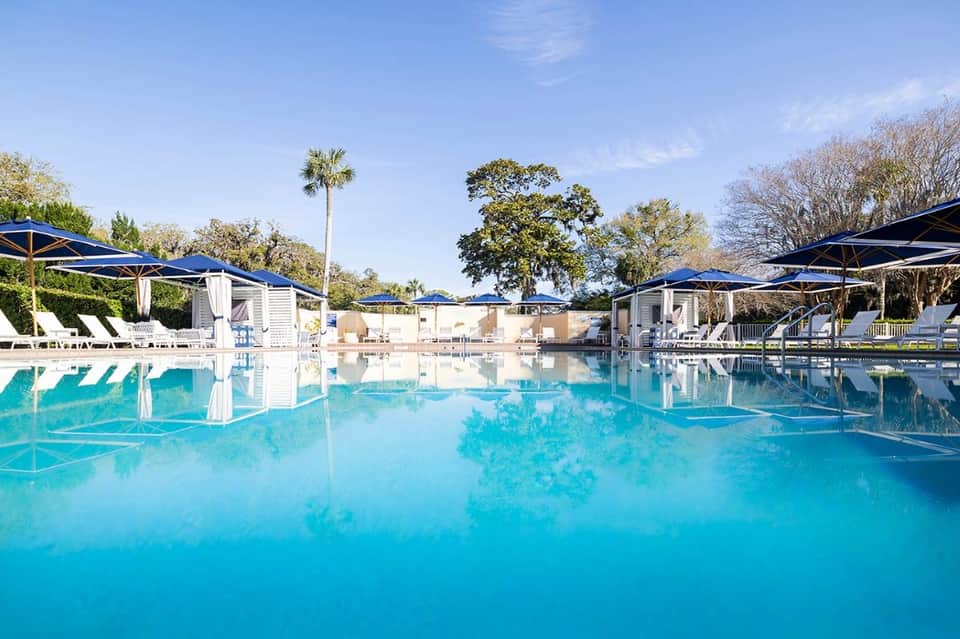 Large resort pool with crystal blue water, blue umbrellas, cabanas, and palm trees
