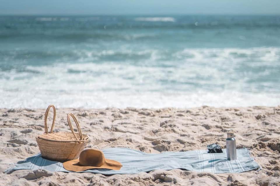 Beach picnic setup with wicker basket, straw hat, and blanket on sand overlooking ocean waves