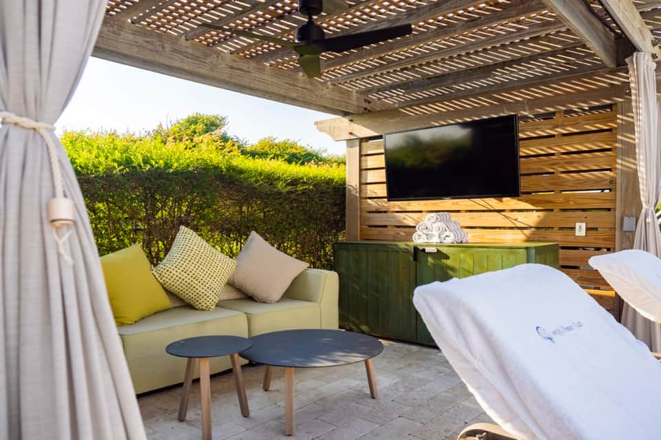 Covered poolside lounge area with pergola, comfortable seating, TV, and lush hedging views