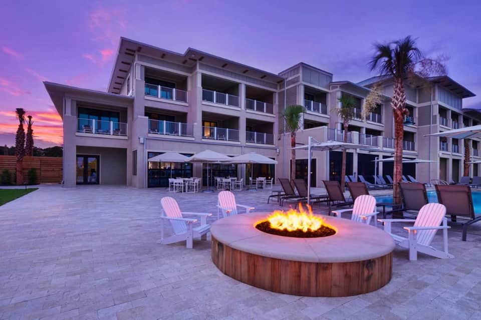 Outdoor firepit lounge at dusk with Adirondack chairs, resort building, and palm trees