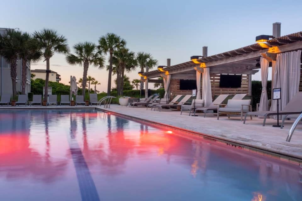 Resort pool at dusk with pink reflection, palm trees, wooden pergola, and lounge areas