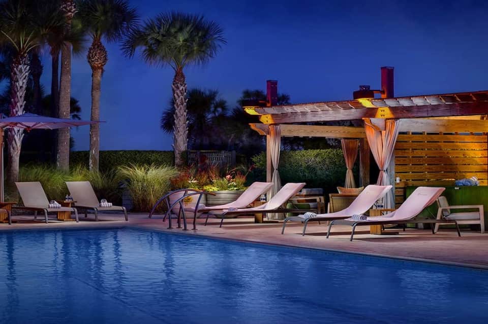 Outdoor pool deck at dusk with lounge chairs, wooden pergolas, palm trees, and ambient lighting