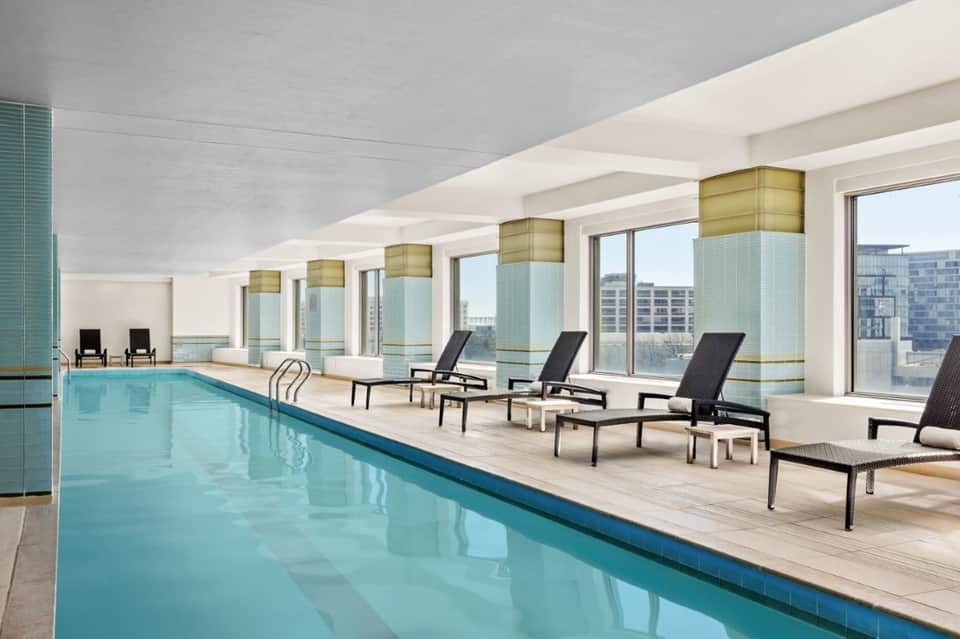 Indoor lap pool with floor-to-ceiling windows, lounge chairs, and city skyline views