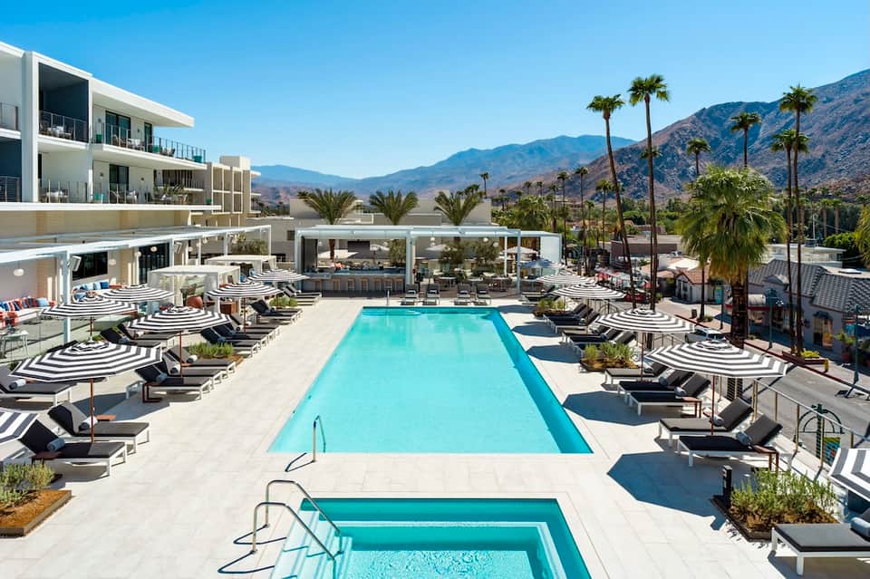 Luxury resort pool with striped umbrellas, mountain views, palm trees, and modern architecture