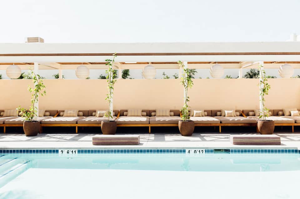 Luxury poolside lounge area with beige cushioned seating, white lanterns, climbing vines, and clear blue pool water
