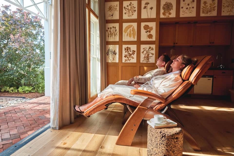 Couple relaxing in wooden lounge chairs indoors with botanical wall art and garden views