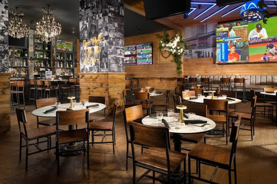 Contemporary sports bar with wooden décor, multiple screens, round tables, chandeliers, and casual dining seating