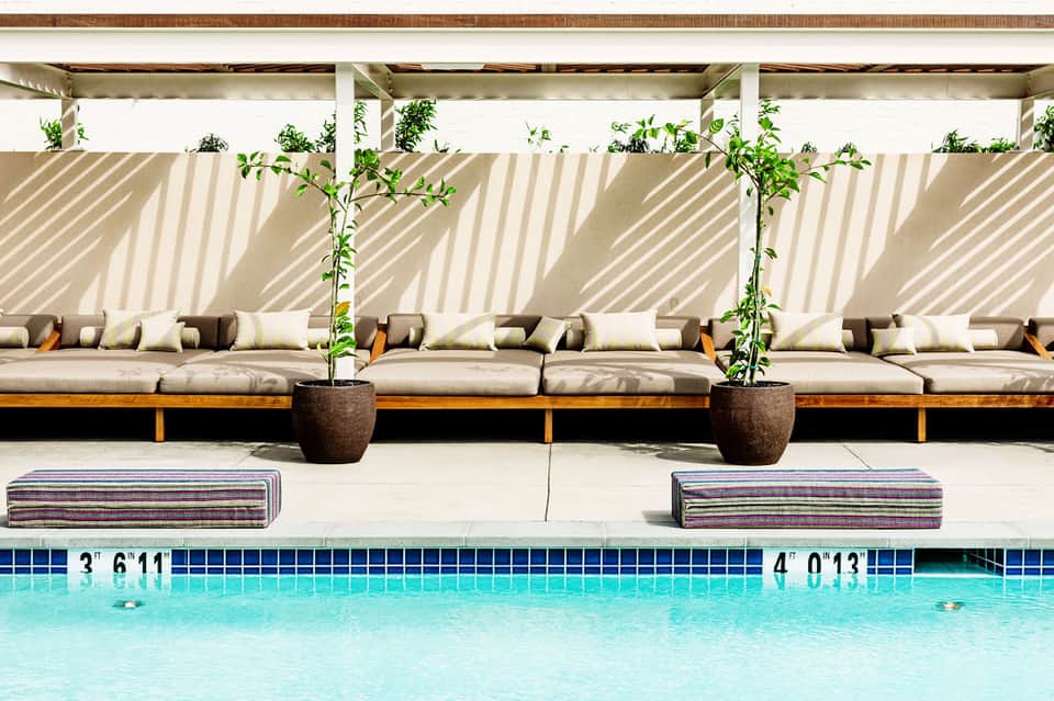 Resort pool with modern lounge seating, pergola shade structure, potted plants, and depth markers on poolside