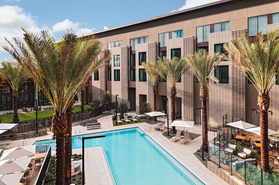Modern hotel courtyard with rectangular swimming pool, palm trees, white umbrellas, and brick building facade