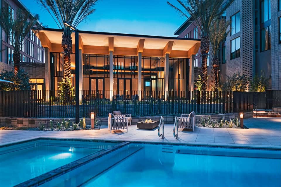 Modern resort pool at night with illuminated pergola, lounge chairs, palm trees, and architectural lighting