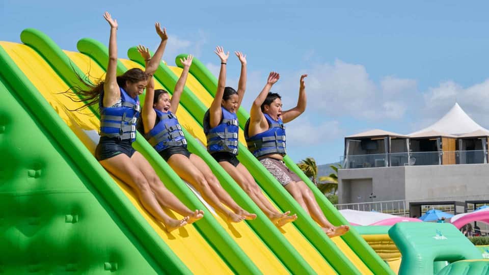 Four people sliding down giant inflatable water slide wearing life jackets with arms raised in joy