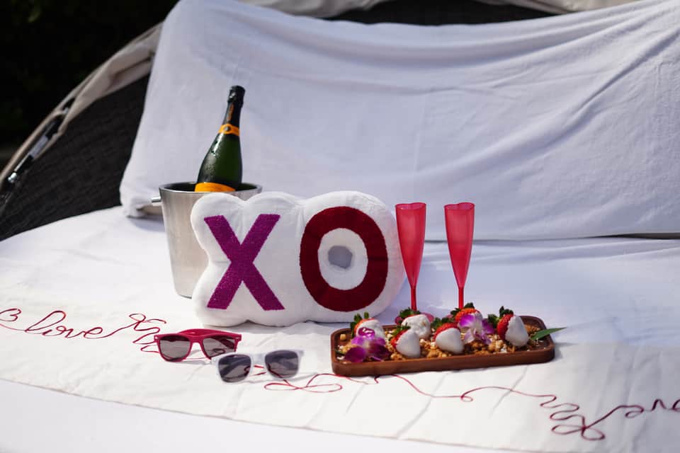 Romantic bed setup with XO pillow decoration, champagne bottle, glasses, and flowers for celebration