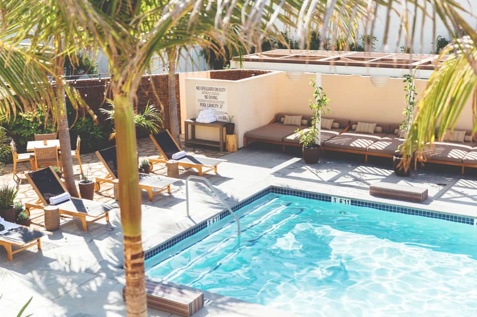 Sunny resort pool deck with palm trees, lounge chairs, and cabanas with shade structures