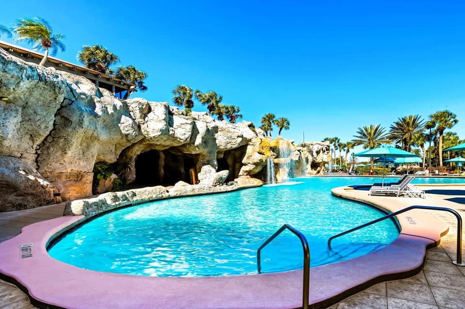 Tropical resort pool with artificial cave feature, rock waterfall, blue umbrellas, palm trees, and sunny sky