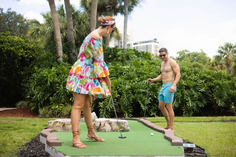 Couple playing mini golf on resort grounds with palm trees and resort buildings in background