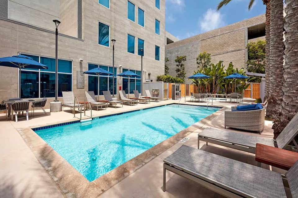Resort pool deck with blue umbrellas, lounge chairs, tropical plants, and modern building