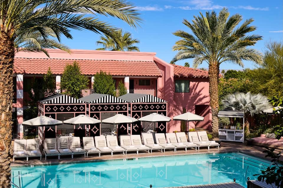 Desert resort poolside with cream lounge chairs, white umbrellas, pink building, and palm trees