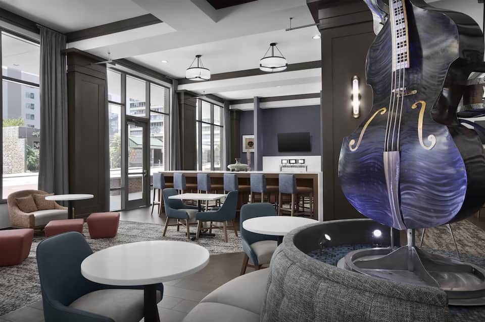 Modern hotel lounge with dark wood accents, red seating, round tables, large windows, bar counter, and decorative cello art