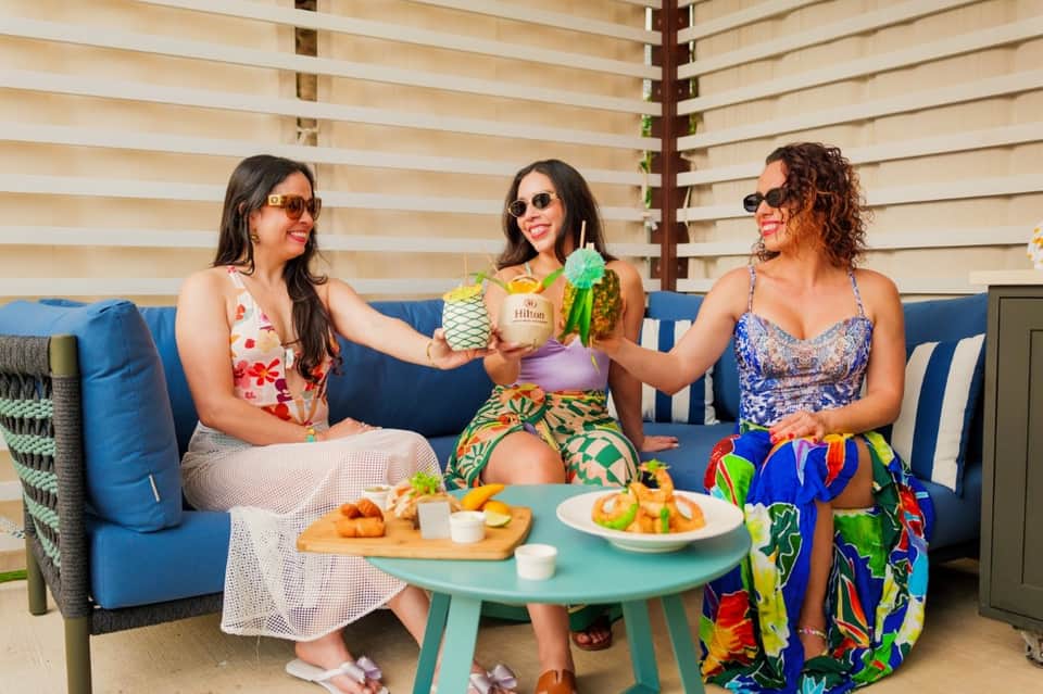 Three women toasting with tropical drinks on hotel terrace with blue couch and wooden privacy screen