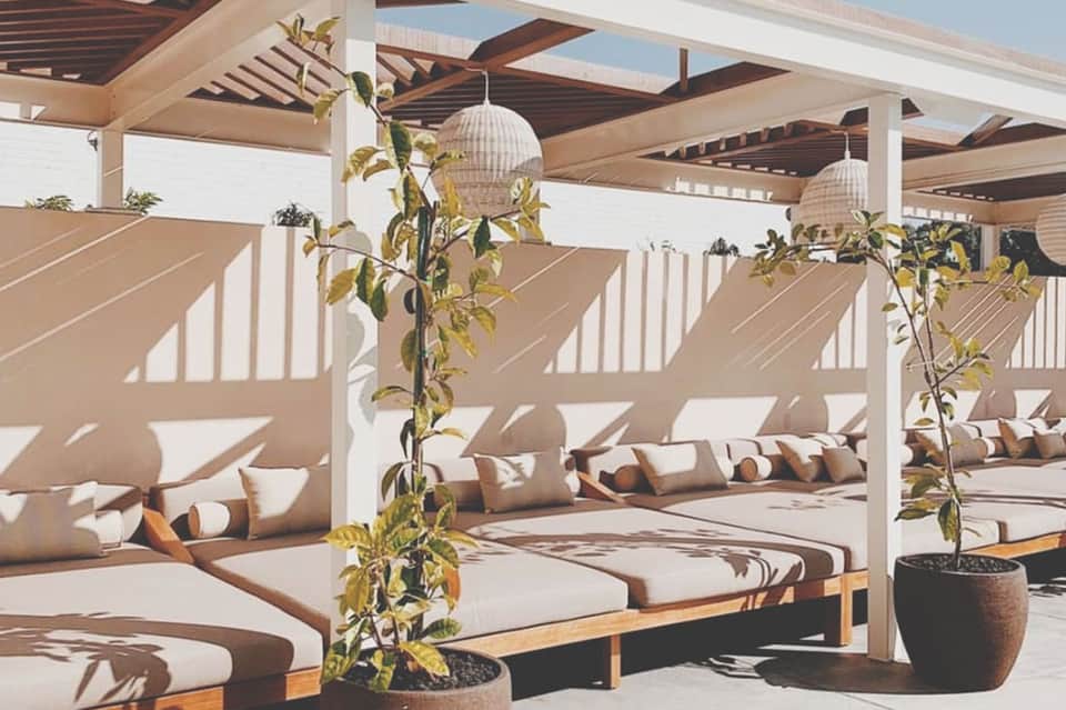 Luxury outdoor lounge area with white pergola, daybed seating, woven pendant lights, and potted plants