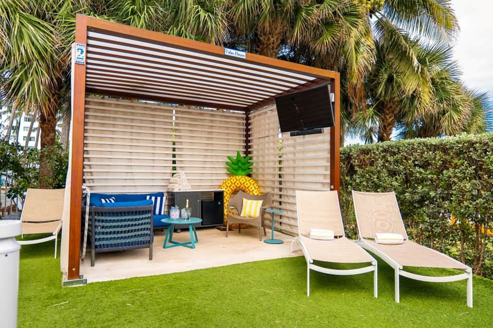 Beachside cabana with wood frame, louvered ceiling, ocean view, blue seating, and adjacent loungers on grass
