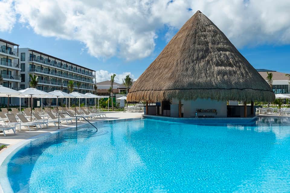 Resort pool area with thatched-roof cabana, lounge chairs, umbrellas, and modern building