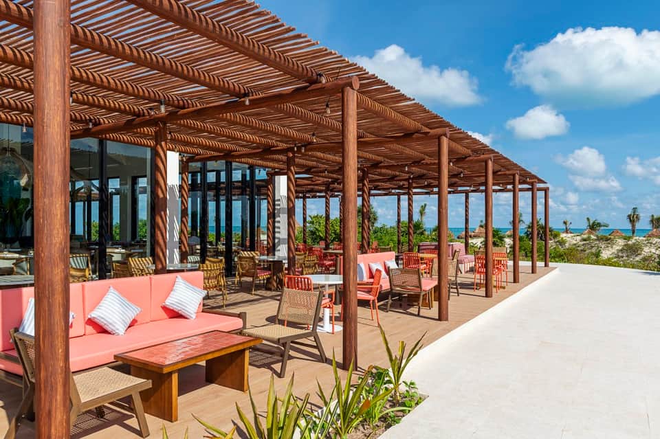 Beachfront dining terrace with wooden pergola, coral seating, ocean views, and palm trees