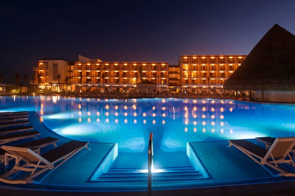 Illuminated resort pool at dusk with warm-lit buildings reflected in glowing blue water