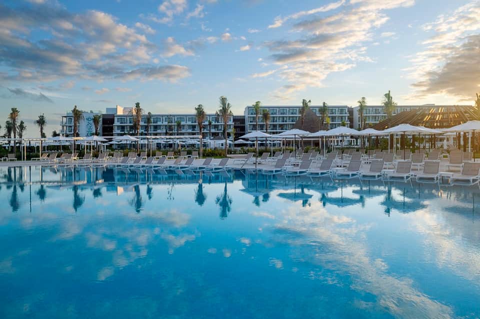 Large resort pool at dusk with white loungers, umbrellas, and modern buildings