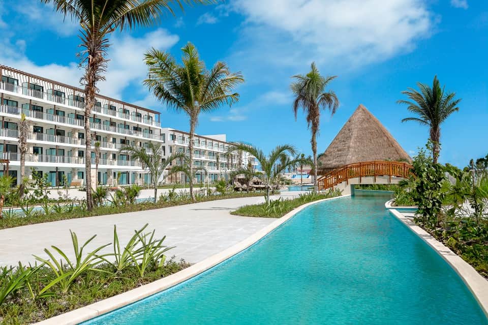 Luxury resort with modern building, turquoise lagoon pool, thatched-roof structure, and palm trees
