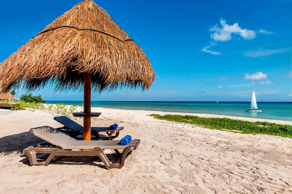 Tropical beach with thatched umbrella, lounge chair, sailboat, and clear turquoise water