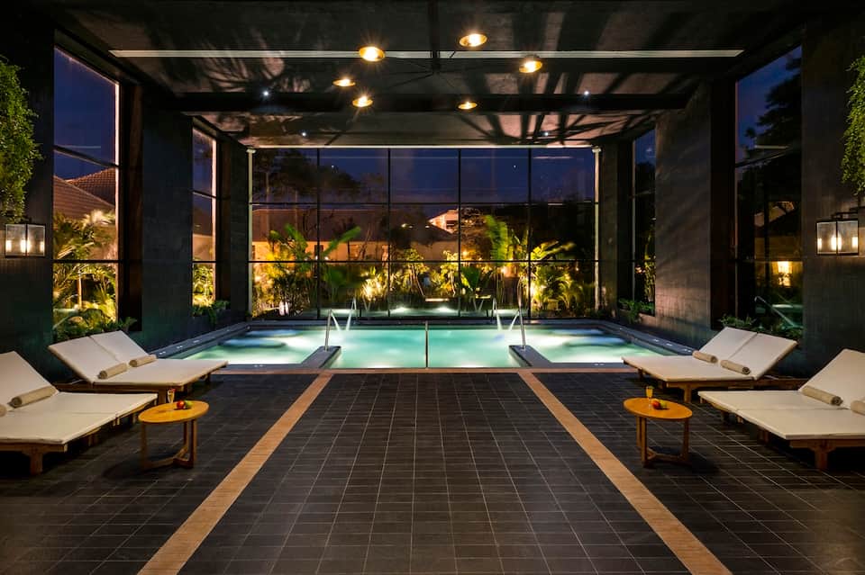 Contemporary indoor pool with glass walls, lounge chairs, ambient lighting, and evening ambiance