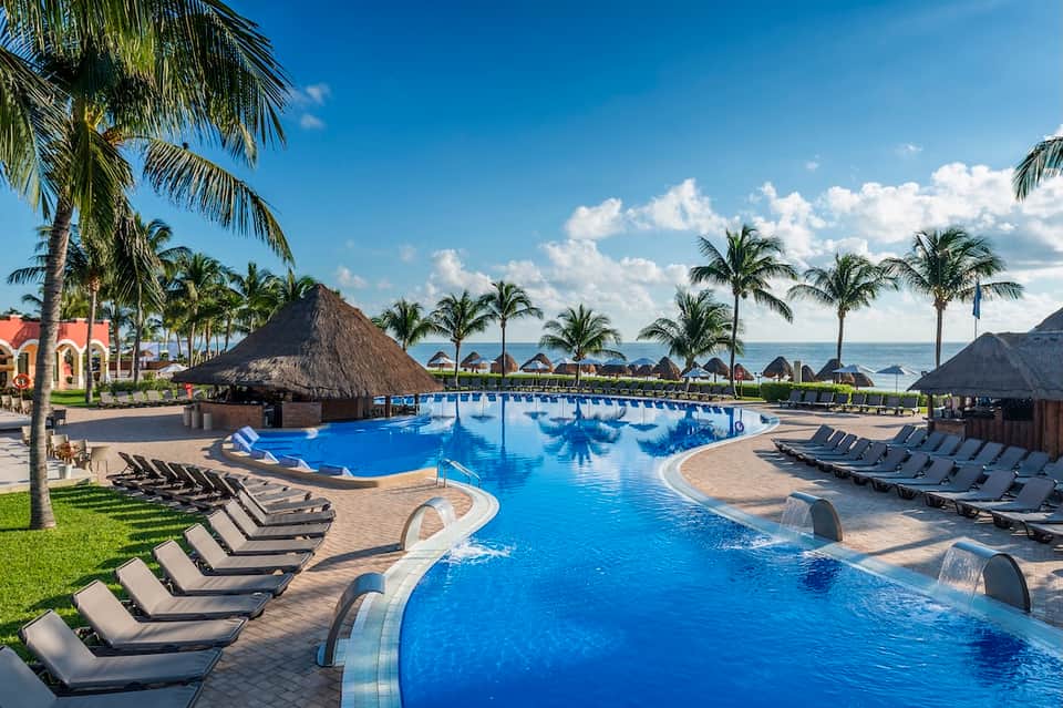 All-inclusive resort pool with palm trees, tiki huts, and ocean views in tropical setting