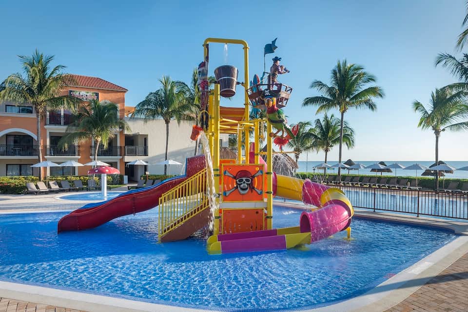 Children's water play area with colorful slide structure in resort pool surrounded by palm trees