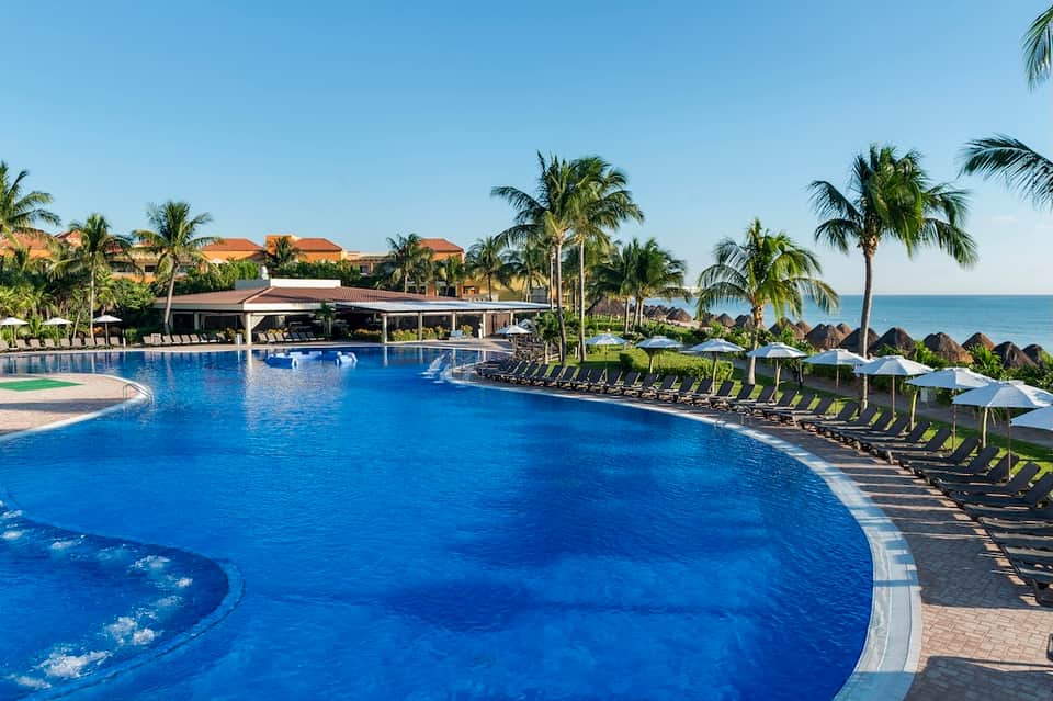 Large resort pool with blue water, palm trees, lounge chairs with umbrellas, and ocean view