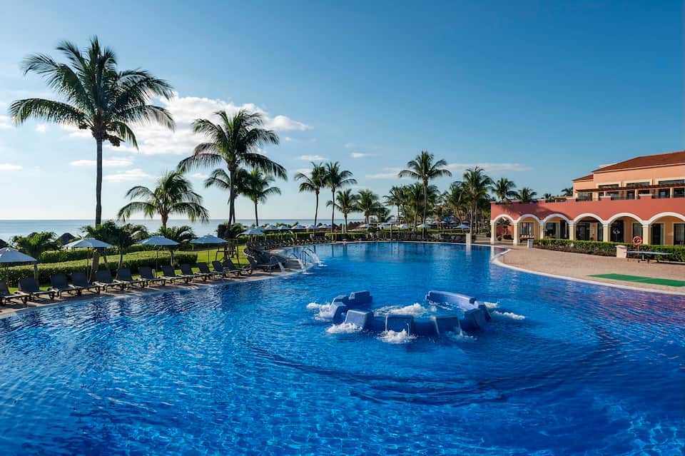 Large resort swimming pool with fountain feature, palm trees, and ocean view beyond