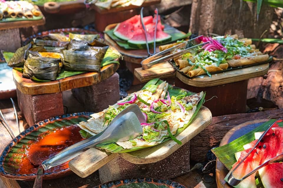 Outdoor tropical buffet display with grilled fish, fresh fruit, salads, and colorful plated dishes
