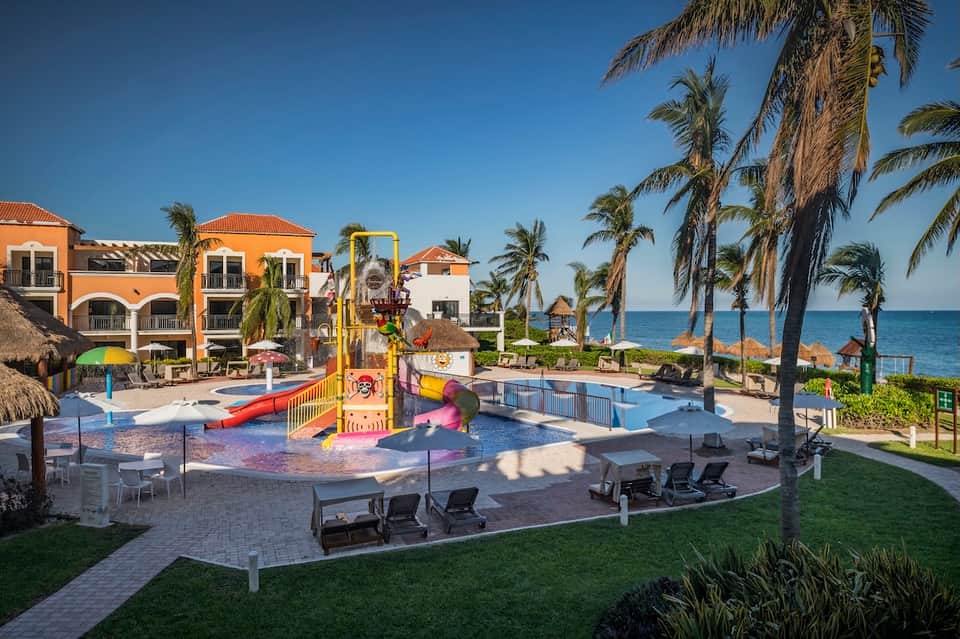 Beachfront resort with colorful buildings, pool, playground, palm trees, and ocean view