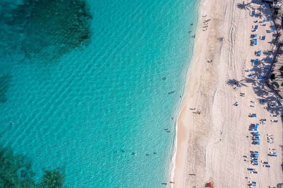 Aerial beach view with turquoise water, white sand, palm tree shadows, and blue lounge chairs