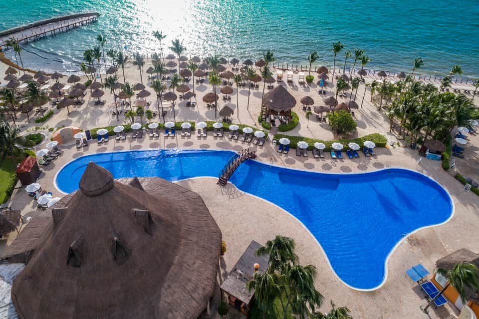 Aerial view of curved resort pool overlooking sandy beach with thatched umbrellas and turquoise ocean