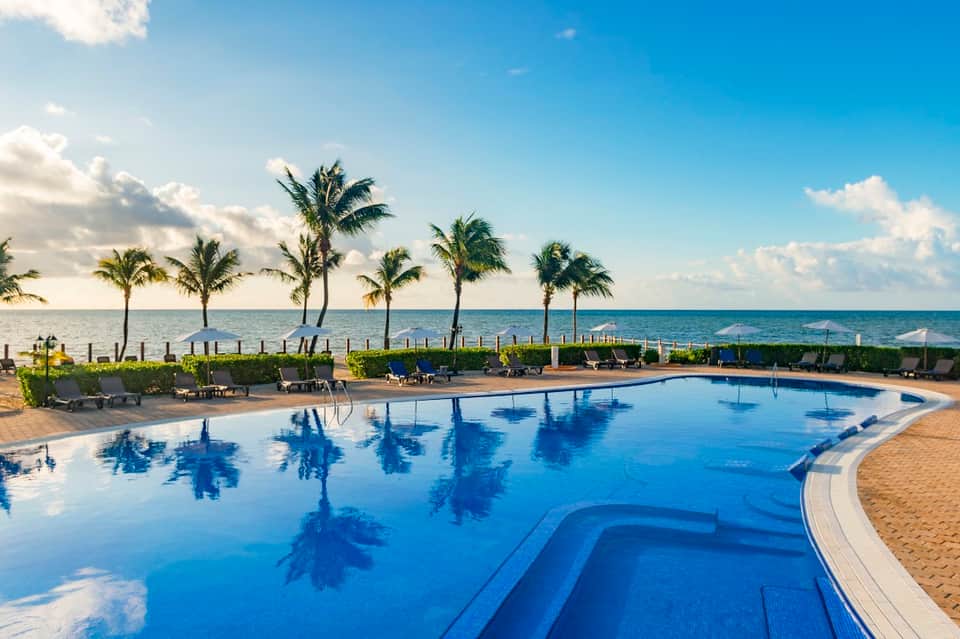 Beachfront resort pool with palm tree reflections, lounge chairs, beach access, and ocean horizon