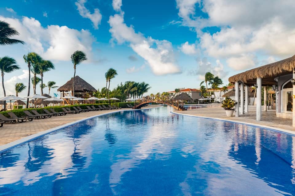 Long resort pool with lounge chairs, palm trees, thatched cabana structures, and manicured grounds under blue sky