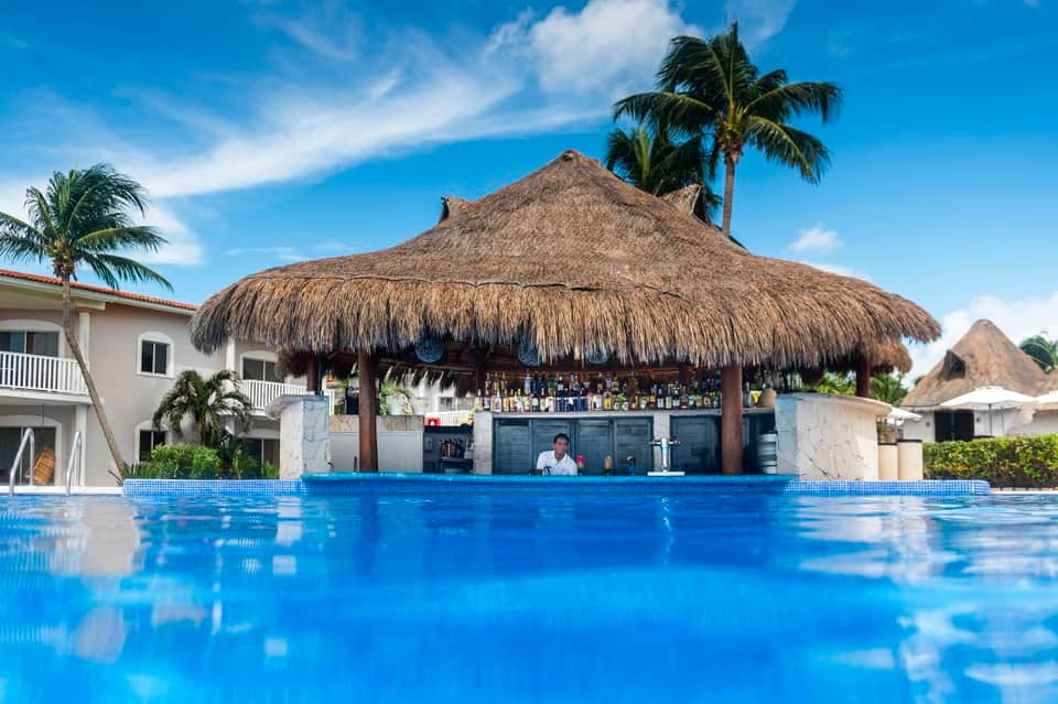Tiki bar with thatched roof beside bright blue pool, palm trees, and resort building