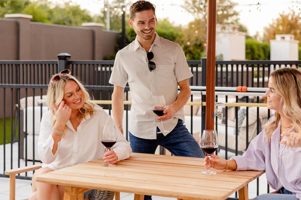 Outdoor patio dining with three people enjoying wine at wooden table, modern railing, and scenic landscape