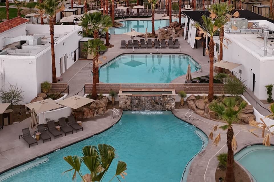 Aerial view of resort pool complex with multiple pools, palm trees, loungers, and buildings