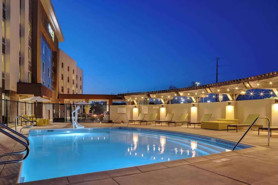 Nighttime resort pool with blue water, lounge chairs, illuminated pergola, and hotel building backdrop